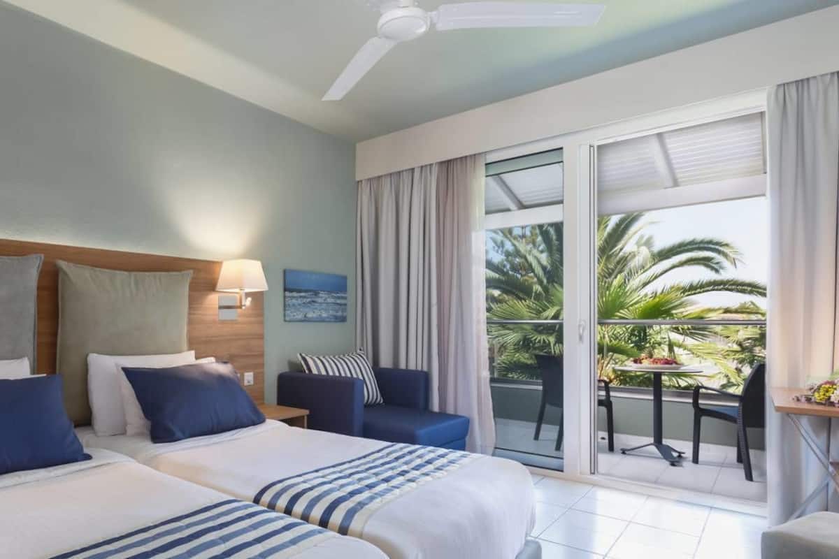 Habitación de hotel con una cama grande, un balcón y vistas a palmeras, además de un ventilador de techo.