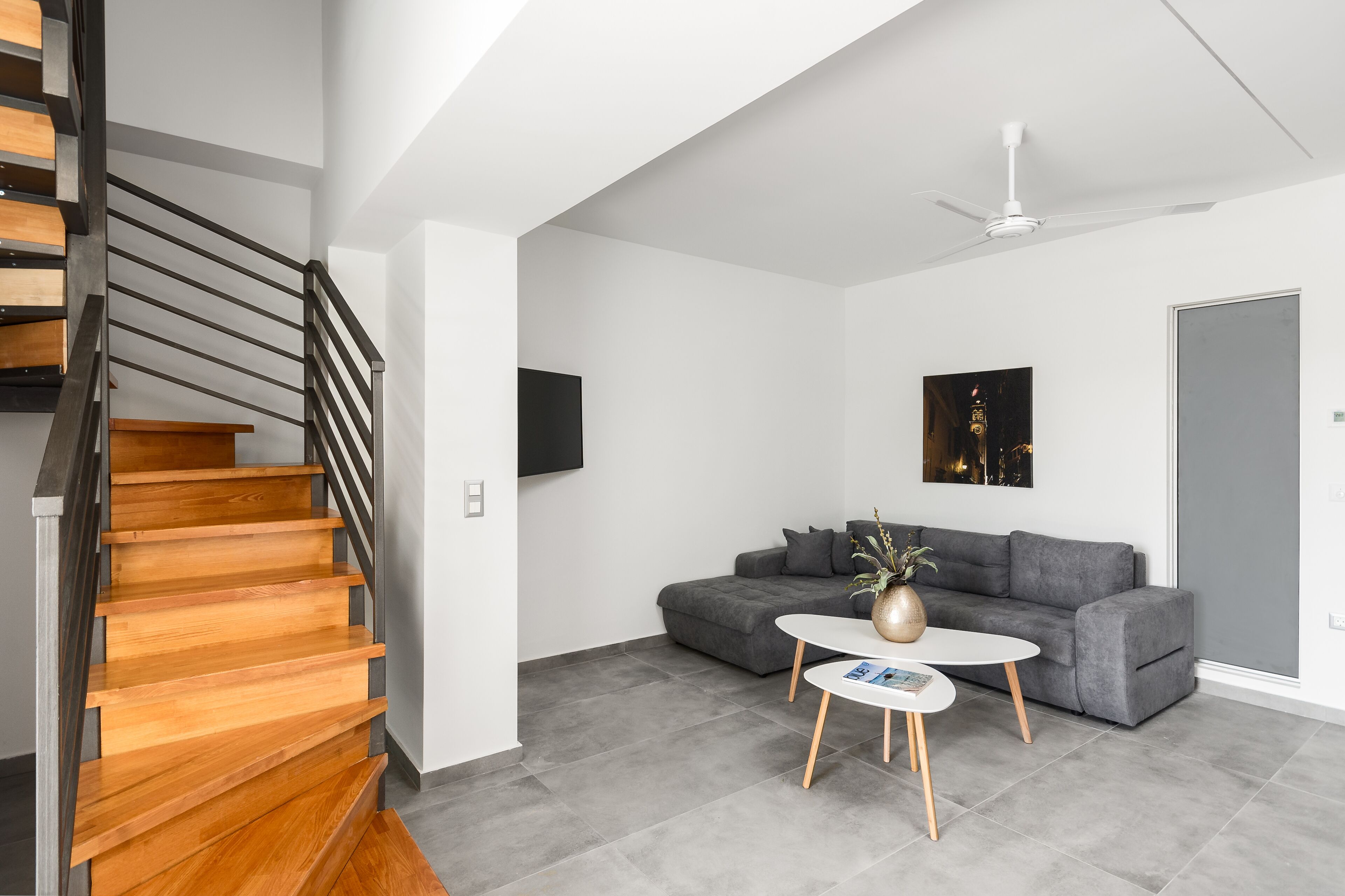 A modern living room with a staircase, grey sofa, white coffee table, and a ceiling fan.
