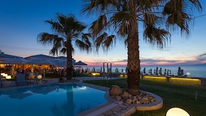 Zona de piscina con palmeras, mobiliario exterior y vista al mar al atardecer.