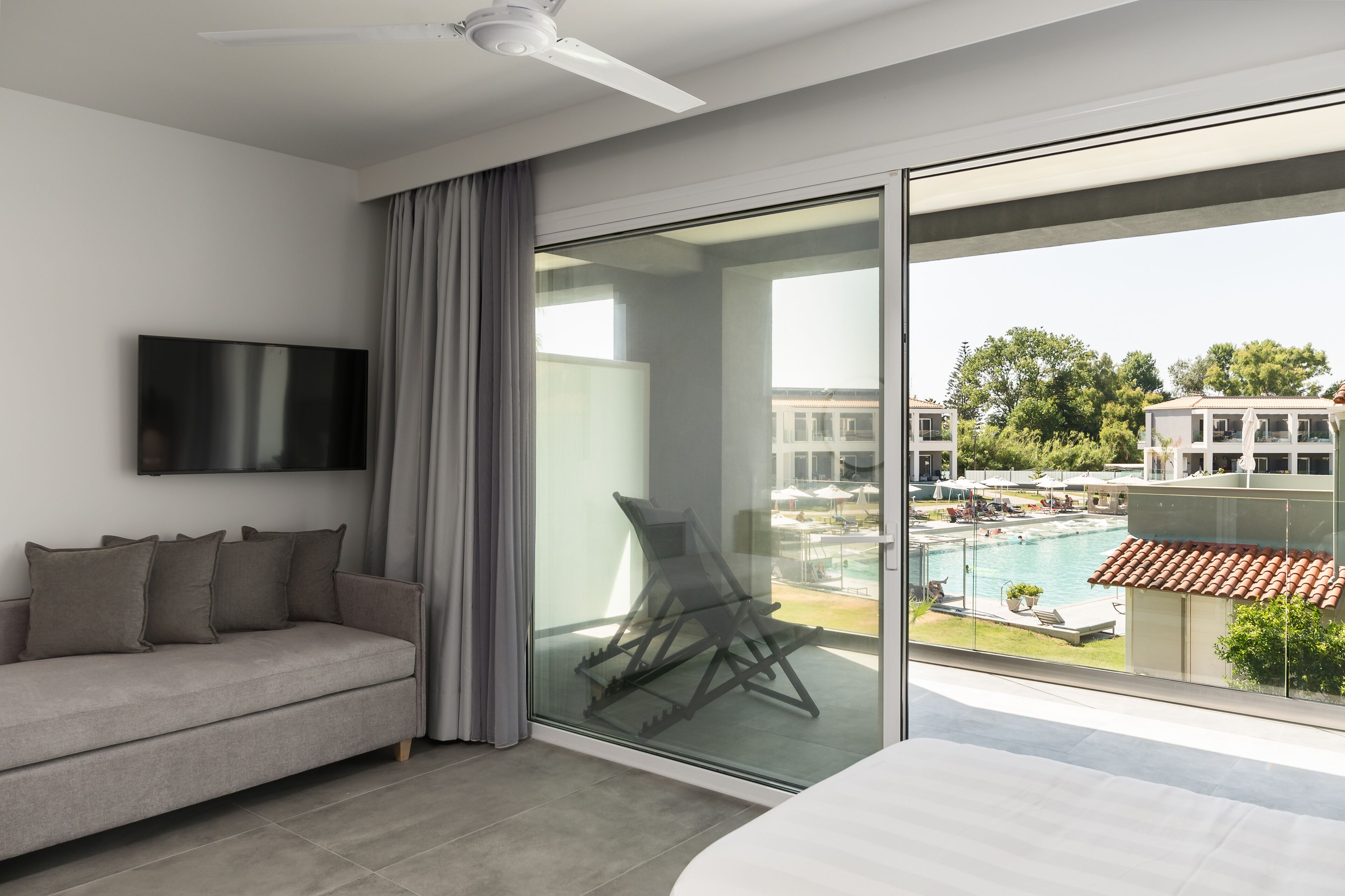A modern hotel room with a sofa, a flat-screen TV, a balcony with a view of a swimming pool and buildings.