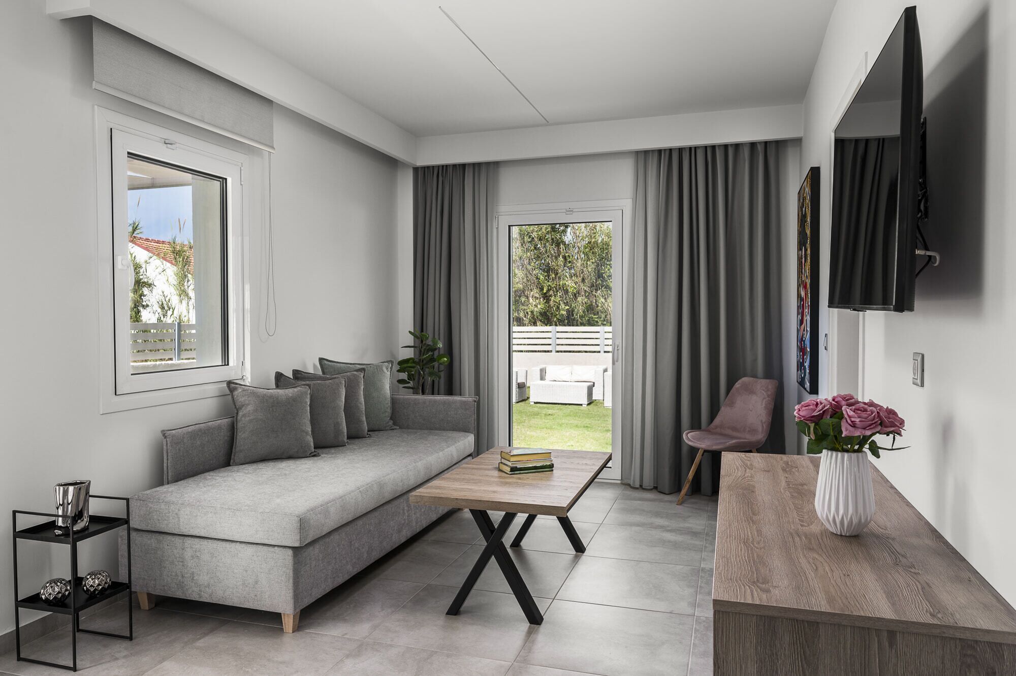 Una sala de estar moderna con un sofá gris, una mesa de centro de madera, un televisor montado en la pared y vistas al jardín a través de una puerta corrediza.