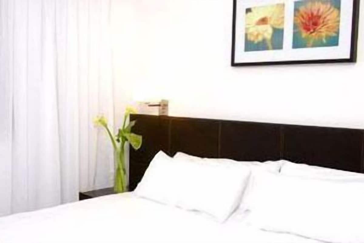 A hotel room with a bed, white curtains, a framed picture on the wall, and a vase with flowers.