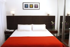 Une chambre d’hôtel avec un grand lit, une table de chevet avec un téléphone, une autre table de chevet avec une lampe, et un tableau encadré au mur.