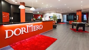Un vestíbulo moderno con un mostrador de recepción rojo identificado con 'DORMERO'. Se observan dos lámparas colgantes, una mesa de billar y una planta en maceta en el centro.