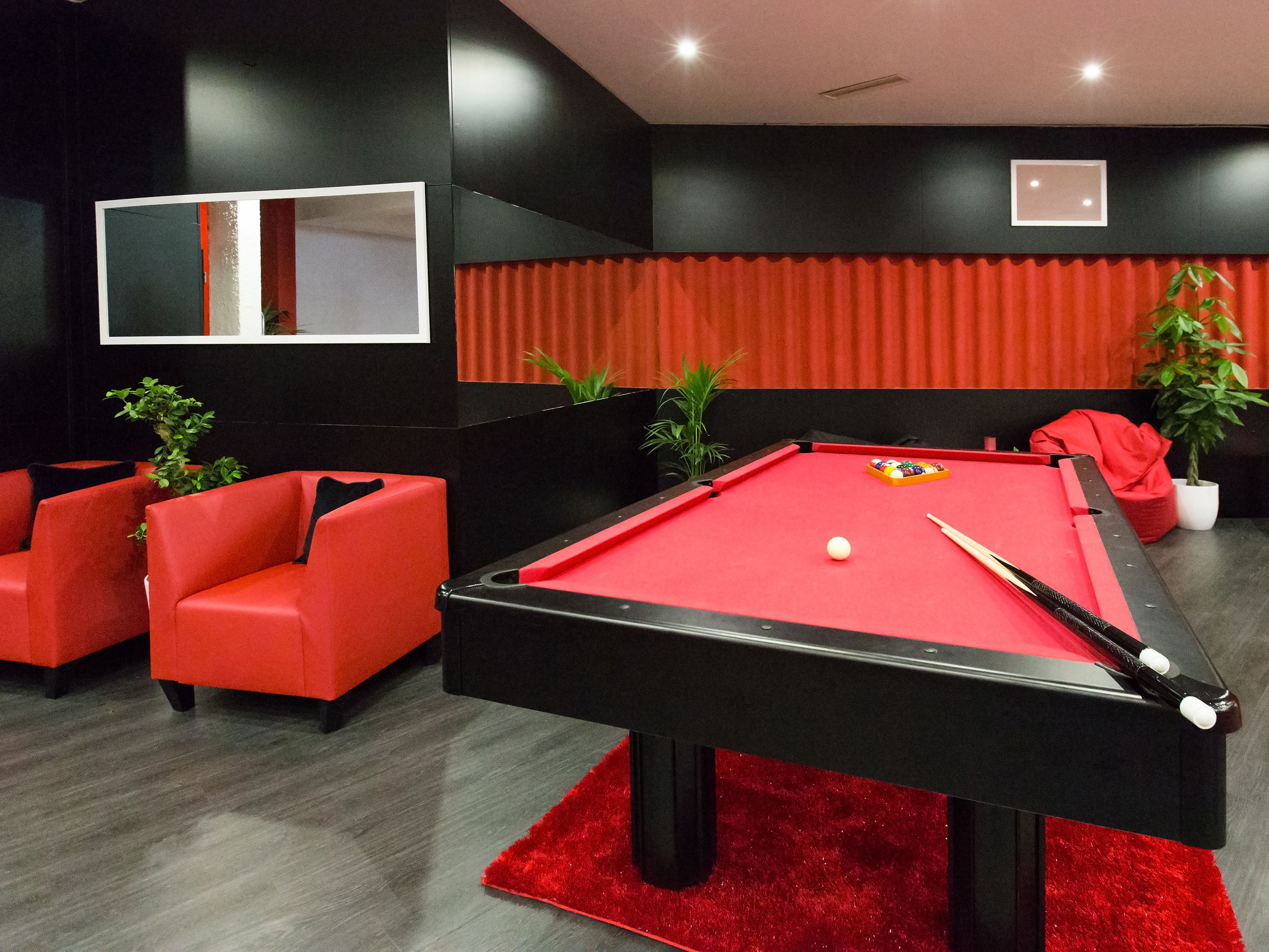 A pool table with a red felt surface, surrounded by red upholstered chairs, in a room with black walls and a red carpet.
