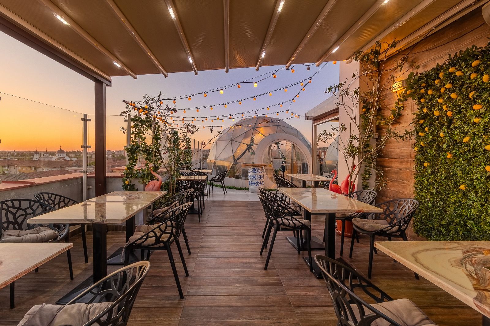 Un espacio de comedor al aire libre con mesas y sillas, una estructura tipo invernadero y vistas a la ciudad.