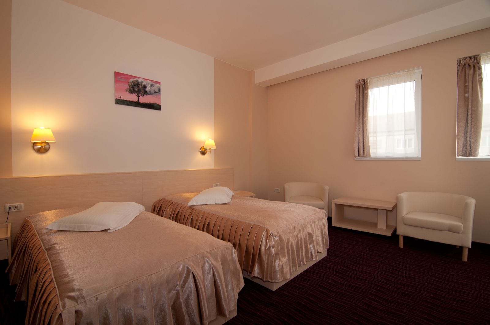 A hotel room with two beds, a chair, a small table, and a painting on the wall.