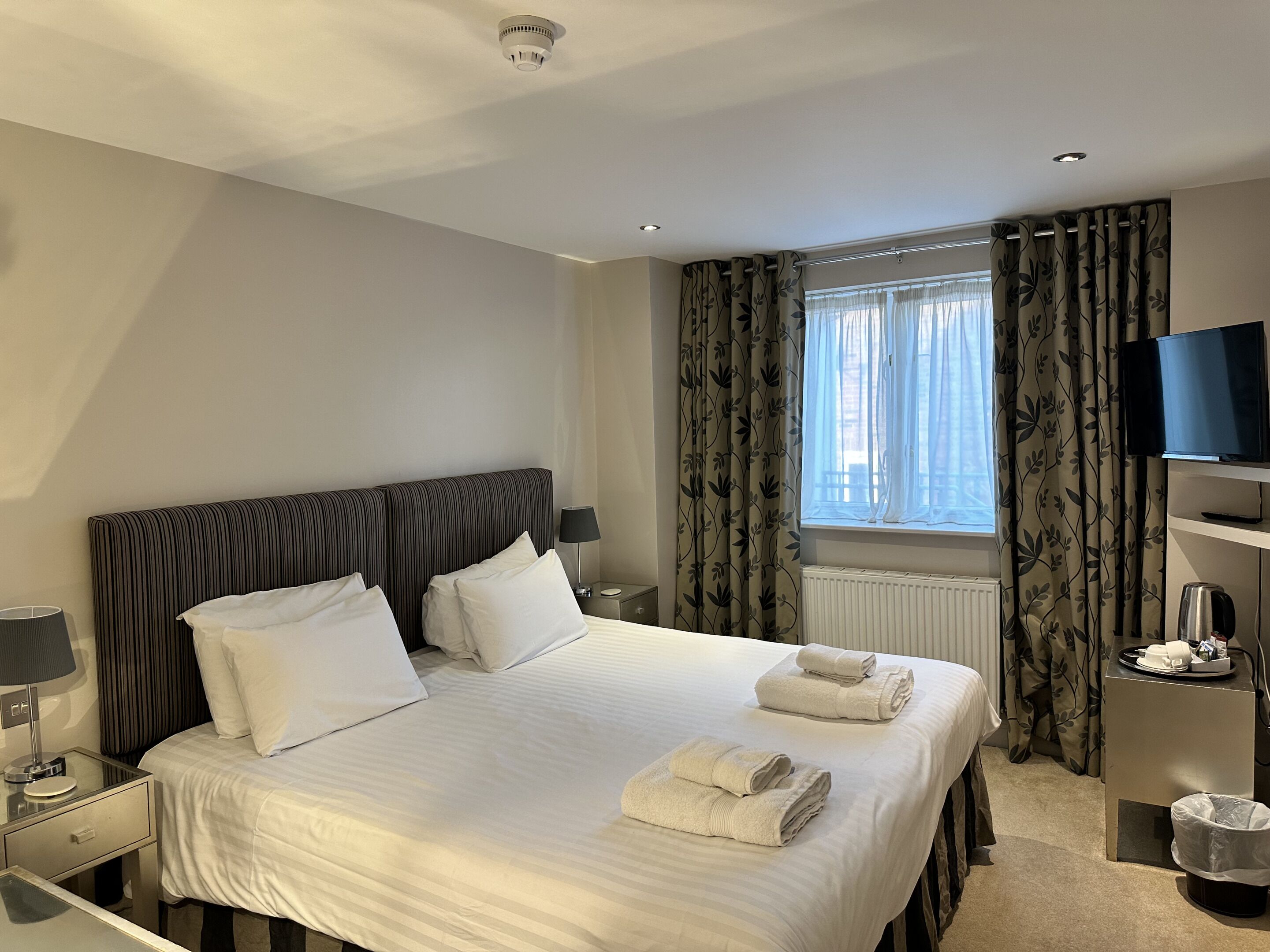A hotel room with a large bed, a TV, a bedside table with a lamp, and a window with curtains.