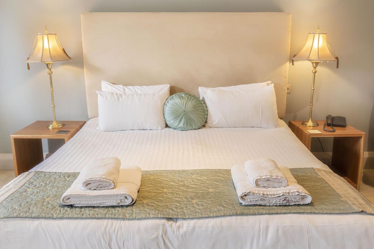 A neatly made bed with white linens, two pillows, a green accent pillow, and two folded towels on a quilted bedspread. There are two bedside tables with lamps and a phone on one of them.