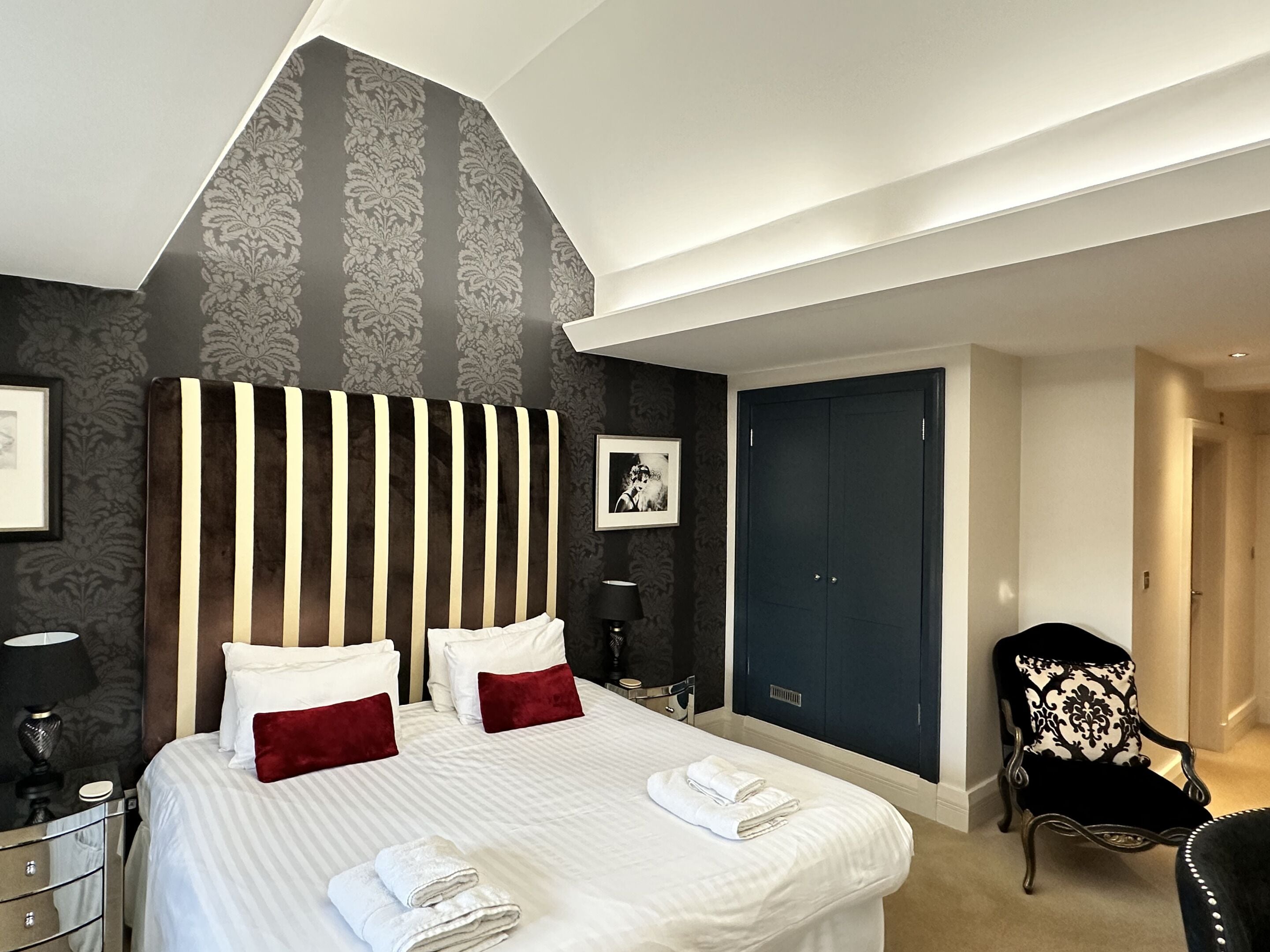 A hotel room with a large bed, a dark wardrobe, a chair, and a desk with a lamp.