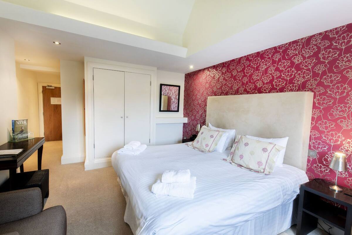 A hotel room with a large bed, white linens, a headboard with a floral pattern, a desk with a chair, and a wardrobe.