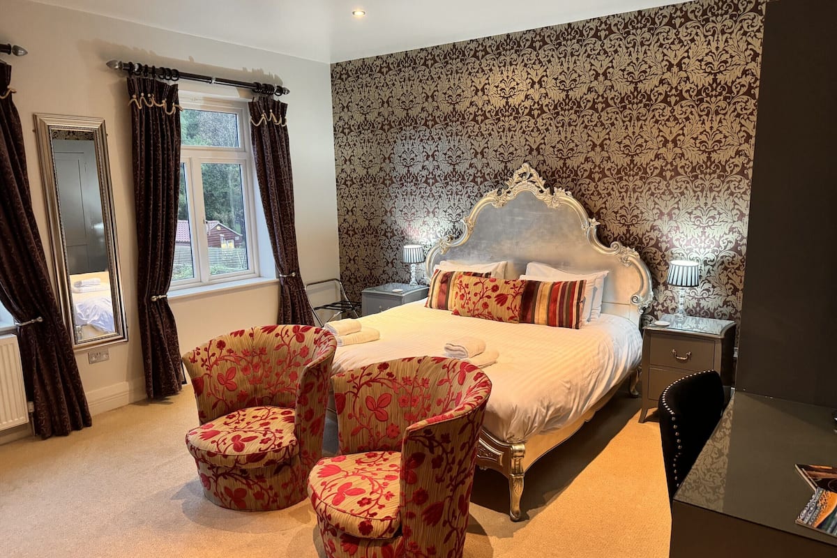 A bedroom with a large bed, two bedside tables, a mirror, and floral-patterned chairs.