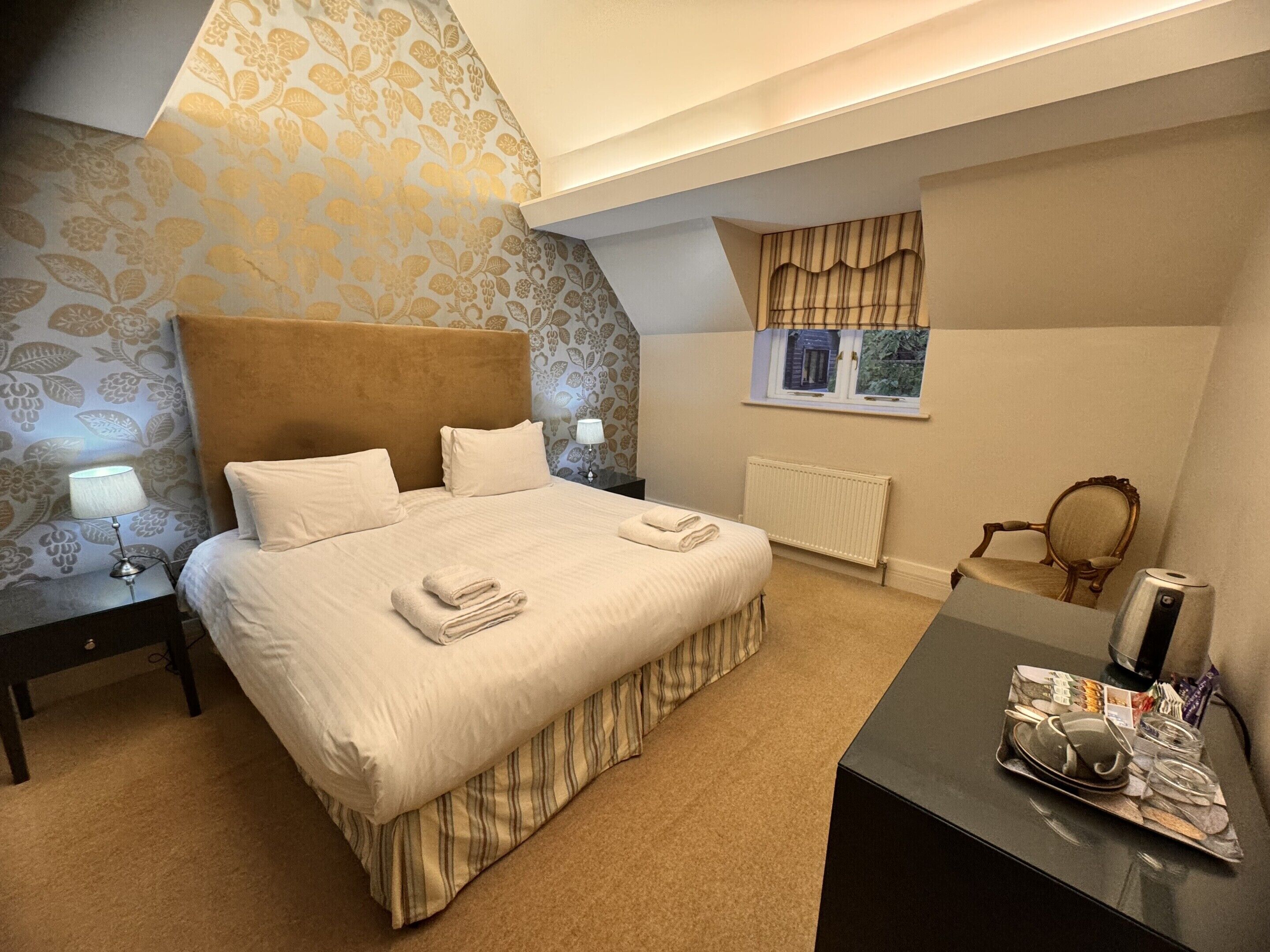A bedroom with a large bed, a bedside table with a lamp, a window with curtains, a radiator, and a small table with a tea set.