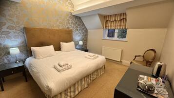 A bedroom with a large bed, a bedside table with a lamp, a window with curtains, a radiator, and a small table with a tea set.