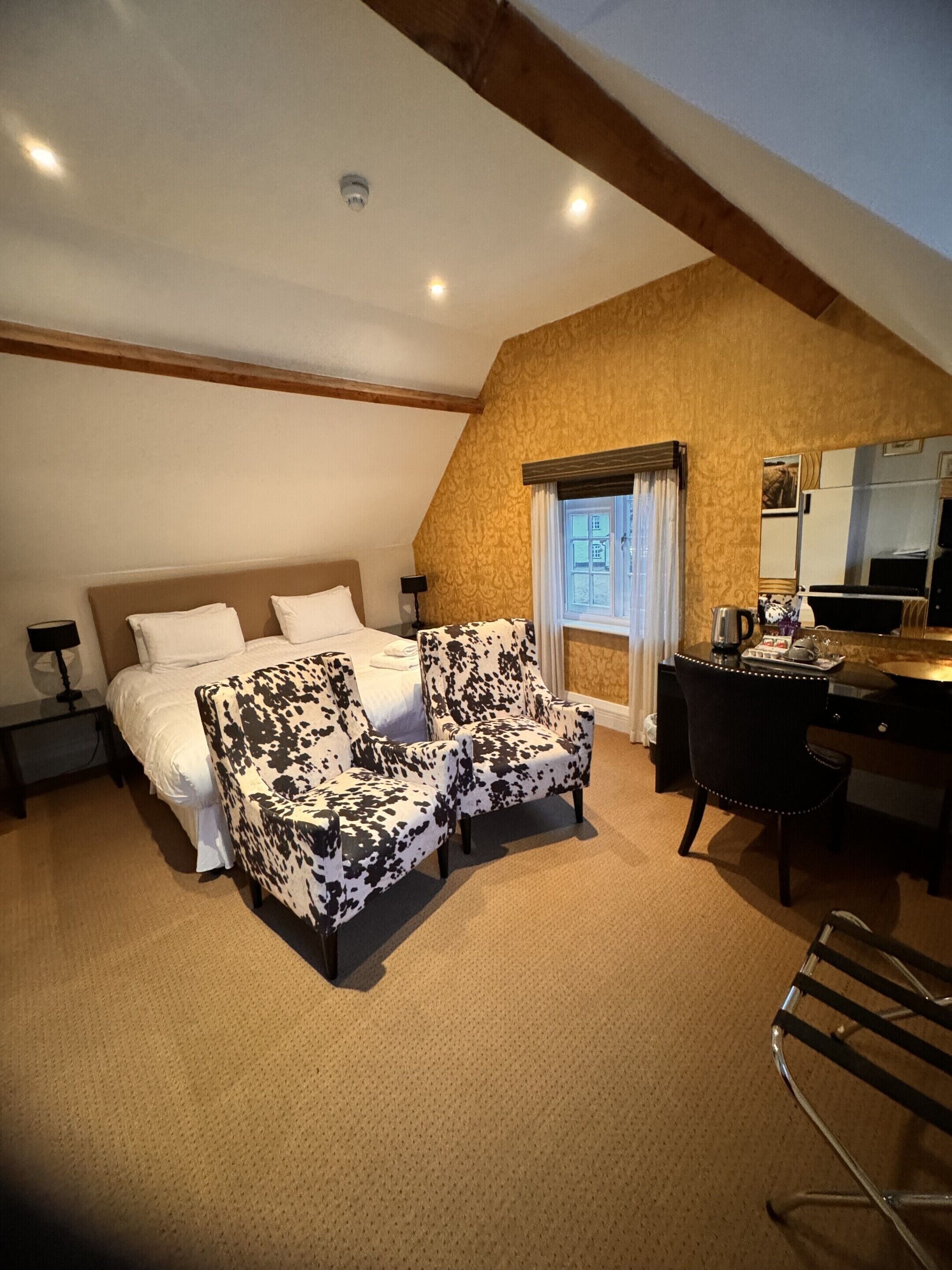 A bedroom with a bed, two armchairs, a desk, a chair, a window, and a mirror.