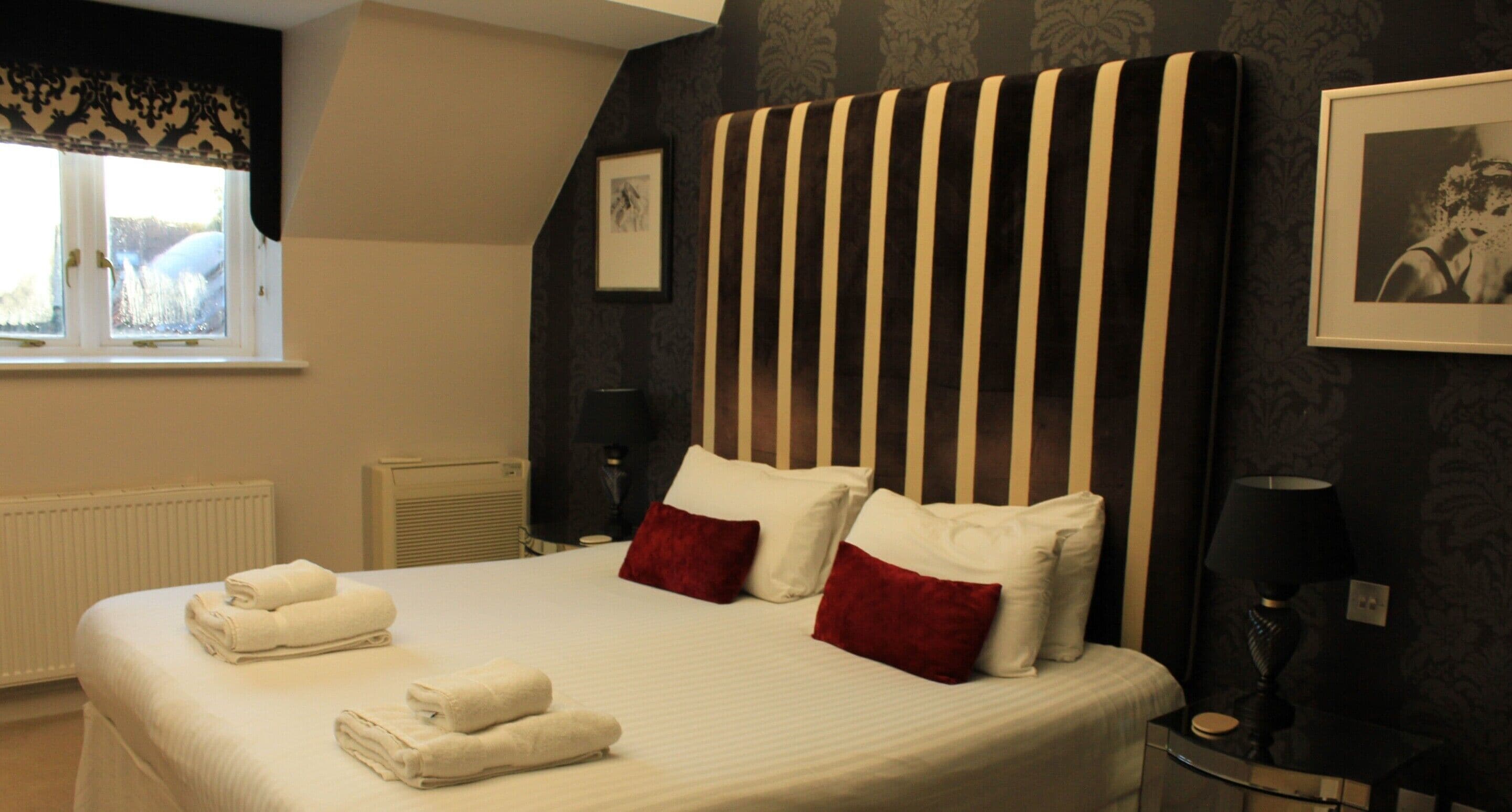 A hotel room with a large bed, two red pillows, a bedside table with a lamp, a framed picture on the wall, and a window with curtains.