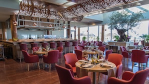 A restaurant with round tables and red chairs, a bar area, and large windows offering a view of the outdoors.