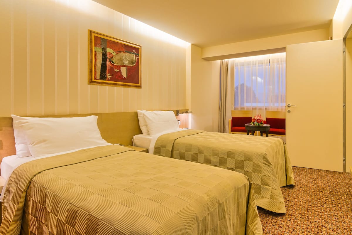 A hotel room with two beds, a painting on the wall, a window with curtains, and a door.