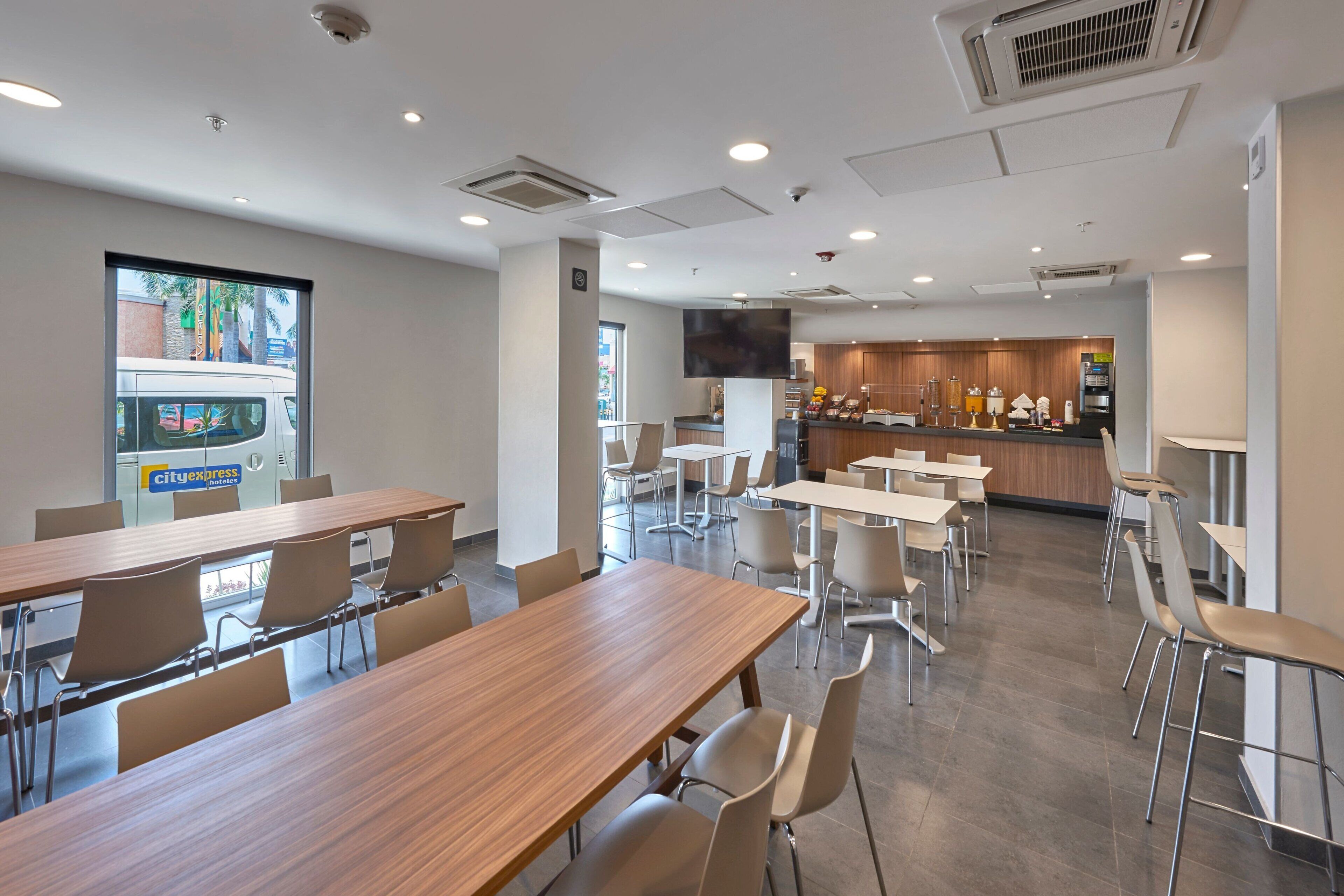 Un comedor moderno con mesas y sillas de madera, una cocina al fondo y una máquina expendedora con el rótulo "cityexpress".