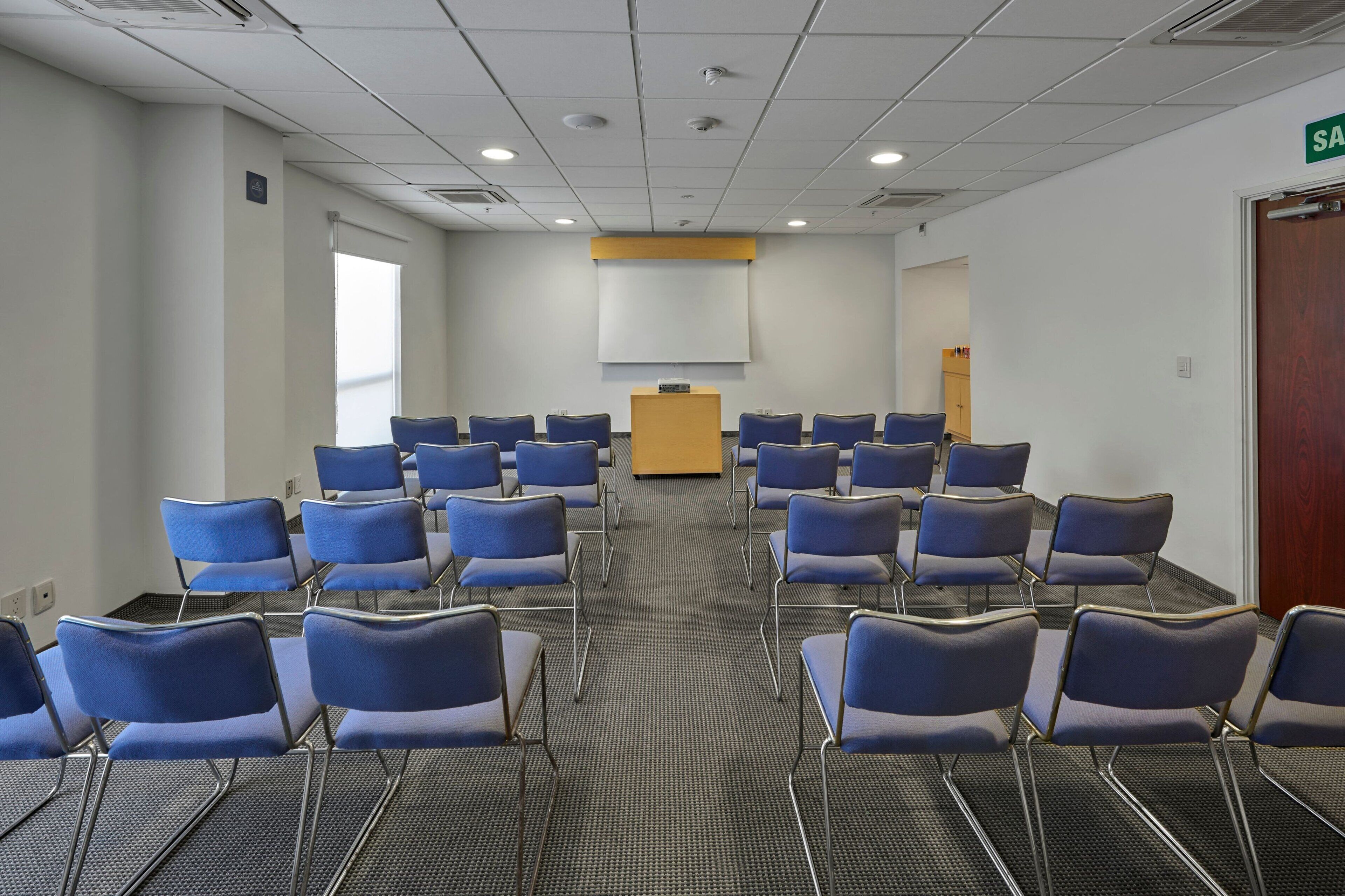 A conference room with rows of blue chairs, a podium, and a projector screen.