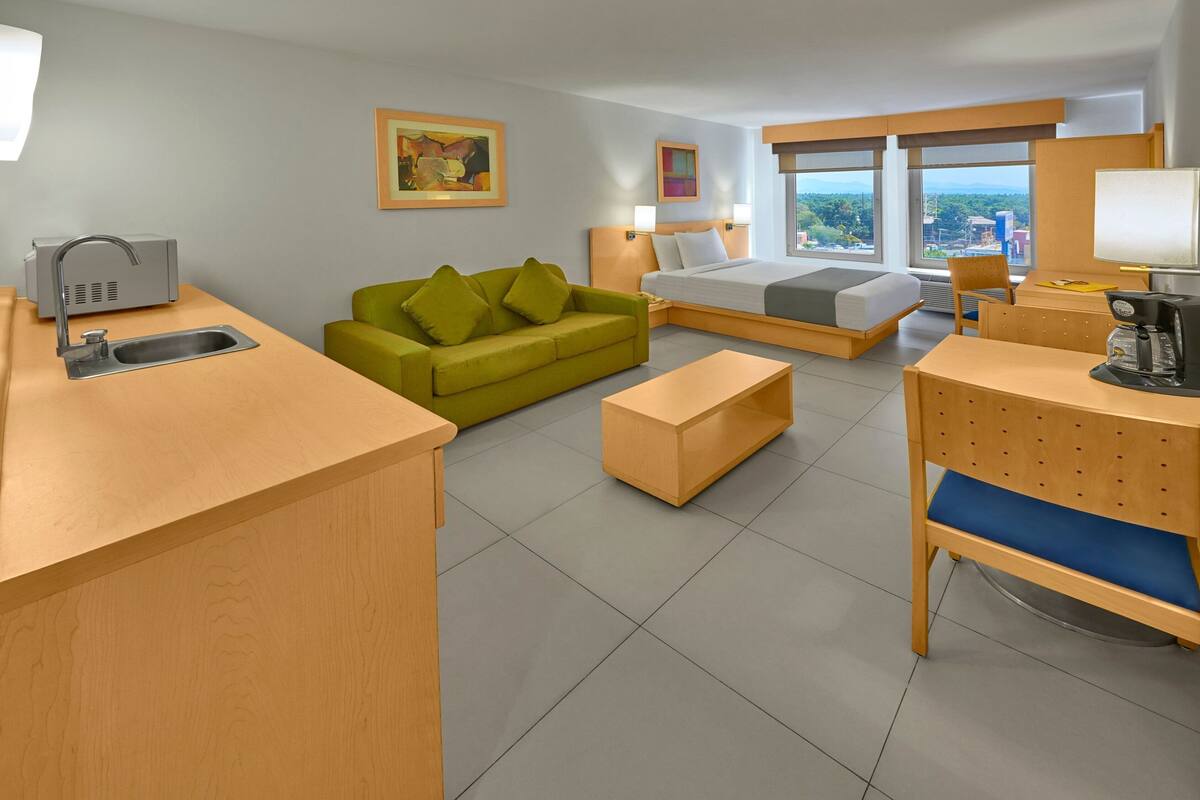 A hotel room with a bed, a green sofa, a wooden desk, a sink, and a view of the city.