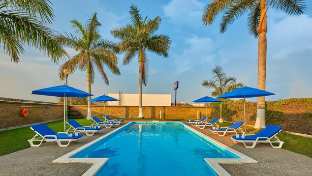 Piscina con sombrillas azules y sillas de descanso, rodeada de palmeras.
