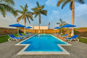 Zwembad met blauwe parasols en ligbedden, omringd door palmbomen.