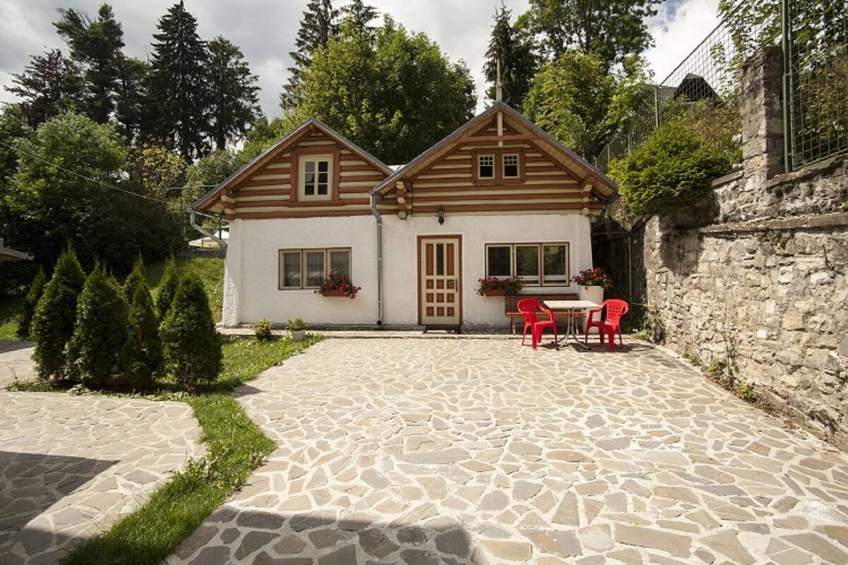 Una casa de dos pisos con un patio empedrado de piedra, fachada de madera y un pequeño jardín con sillas rojas y plantas en macetas.