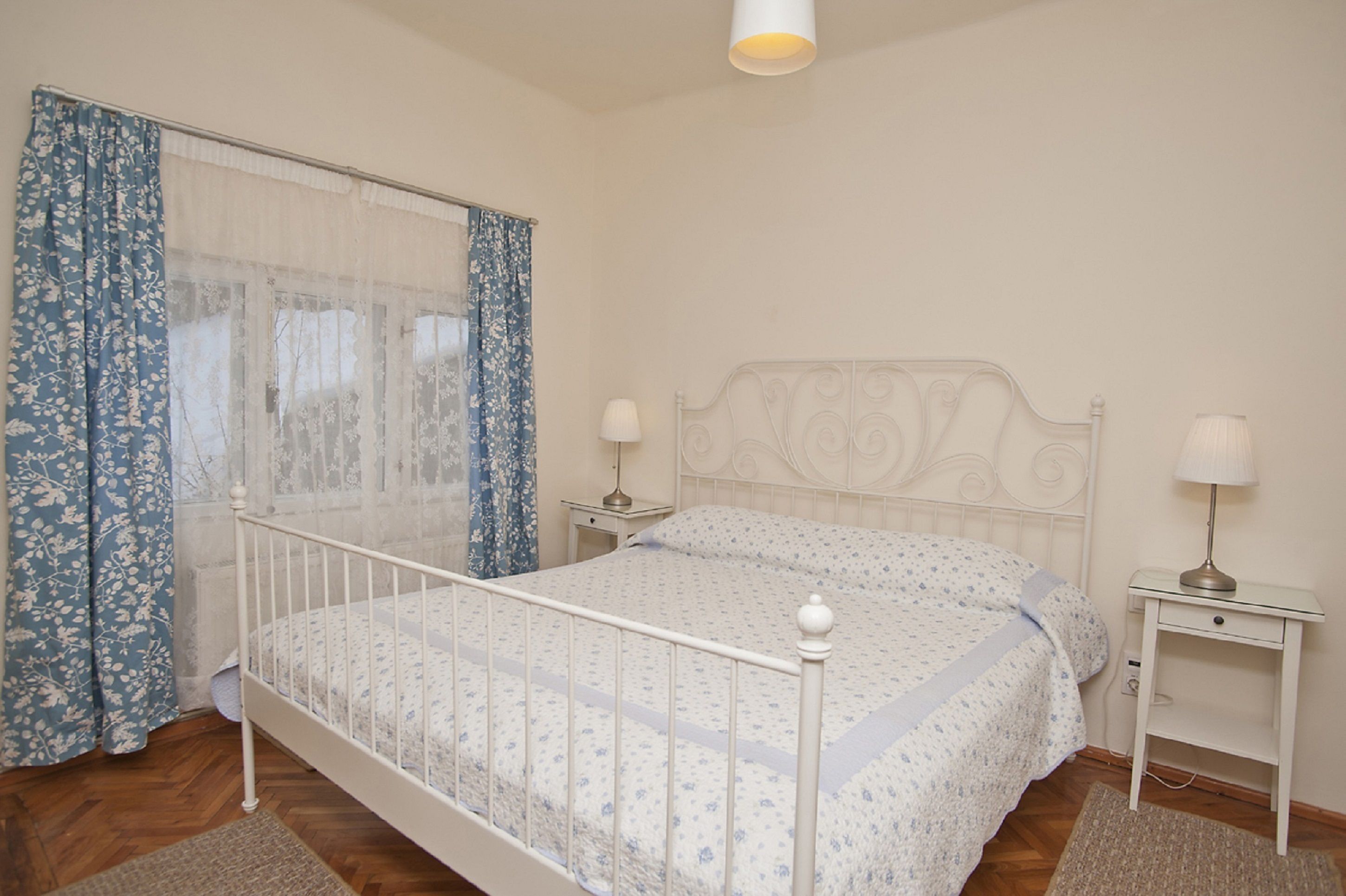 Un dormitorio con estructura de cama metálica blanca, ropa de cama con motivos florales, dos mesitas de noche con lámparas y una ventana con cortinas azules.