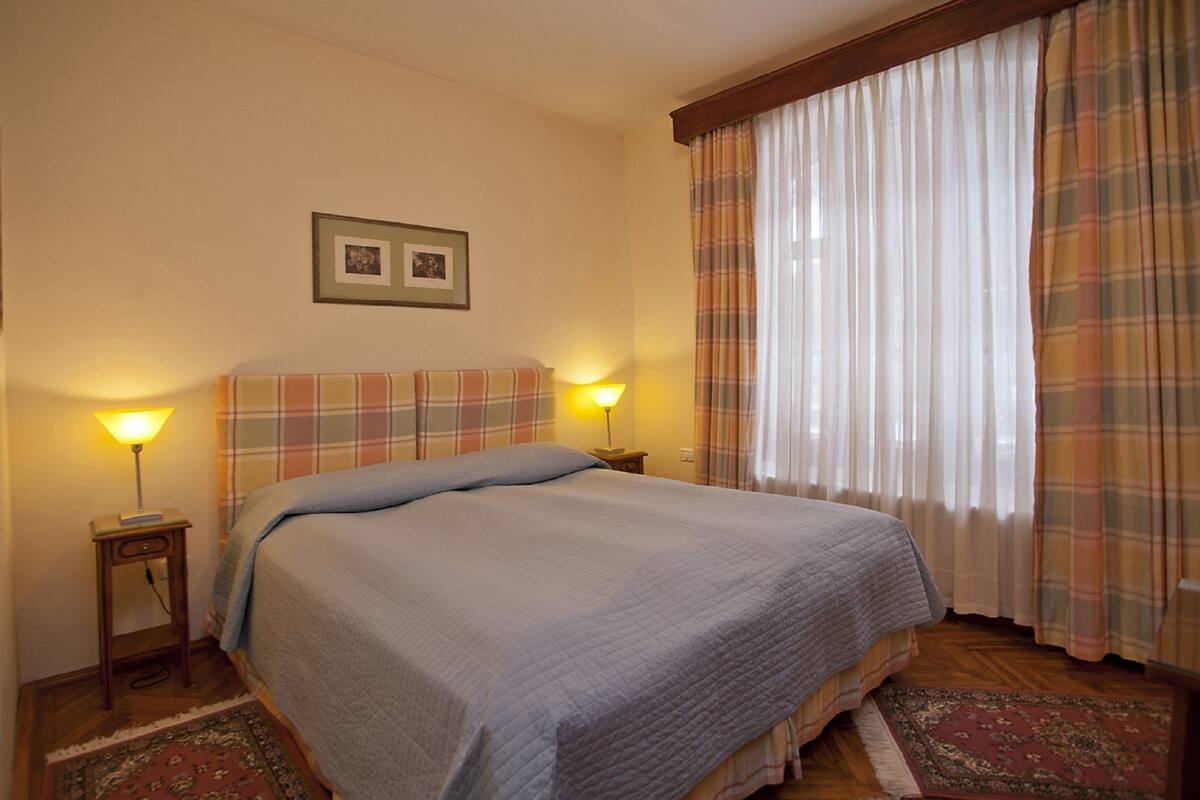 Un dormitorio con una cama, dos mesitas de noche con lámparas, un cuadro en la pared y cortinas a cuadros.