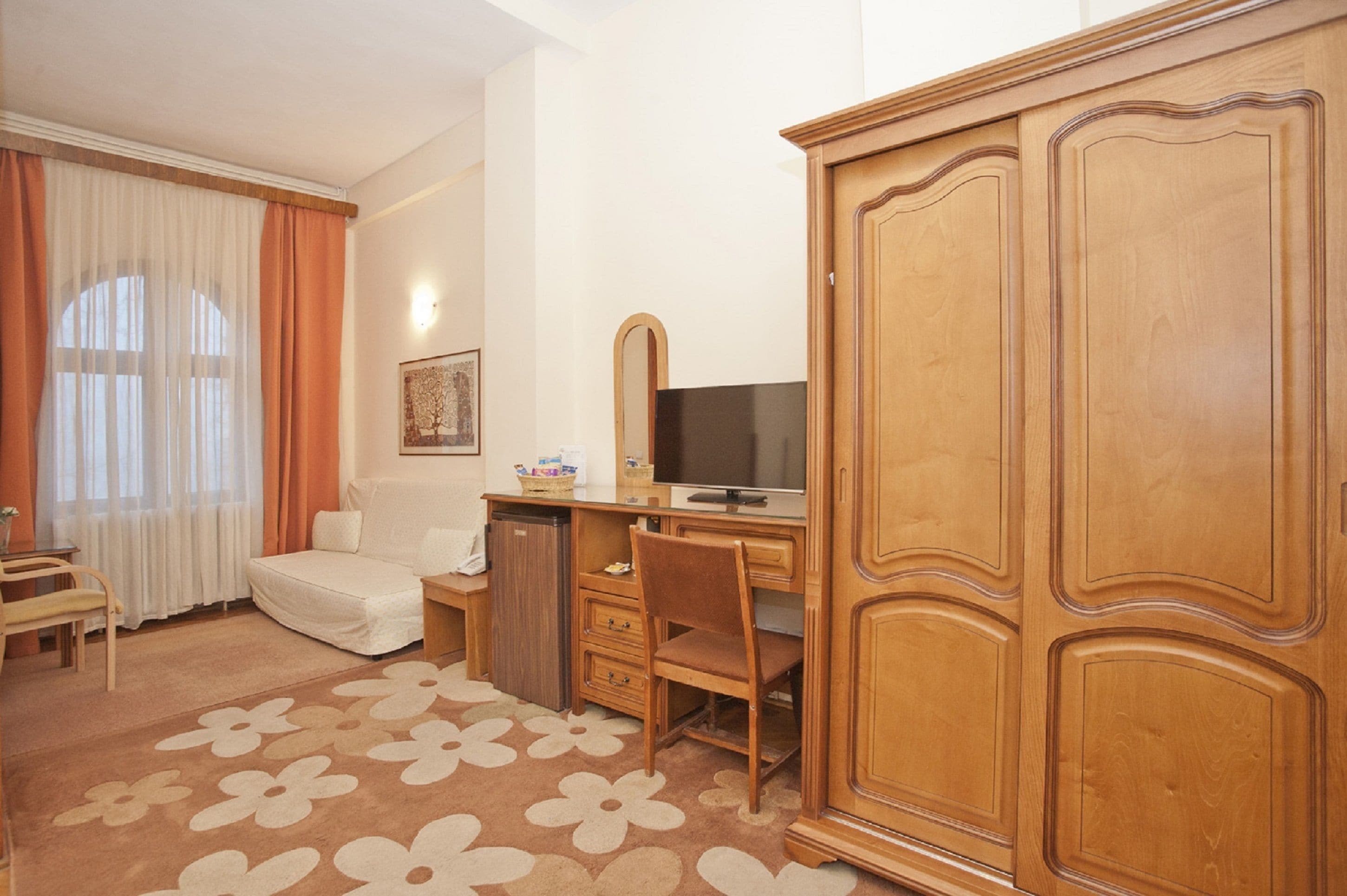 A hotel room with a wooden wardrobe, a desk with a computer, a chair, a sofa, and a small table with a lamp.