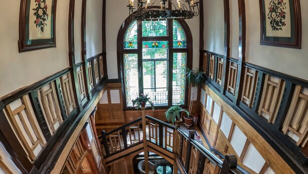 Un magnifique escalier en bois, agrémenté de rampes et surmonté d’un lustre, mène à un niveau inférieur où se trouvent un espace salon et une grande fenêtre à vitraux.