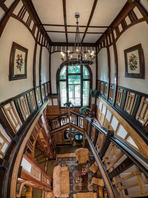 Un magnifique escalier en bois, agrémenté de rampes et surmonté d’un lustre, mène à un niveau inférieur où se trouvent un espace salon et une grande fenêtre à vitraux.