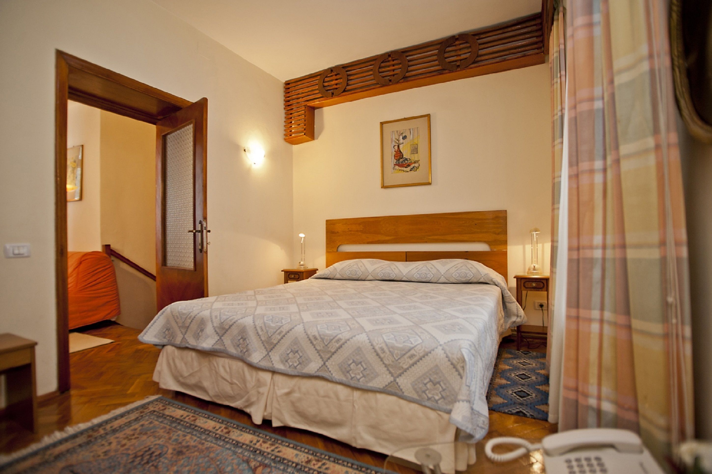 A bedroom with a wooden headboard, a patterned bedspread, a nightstand with a lamp, and a telephone.