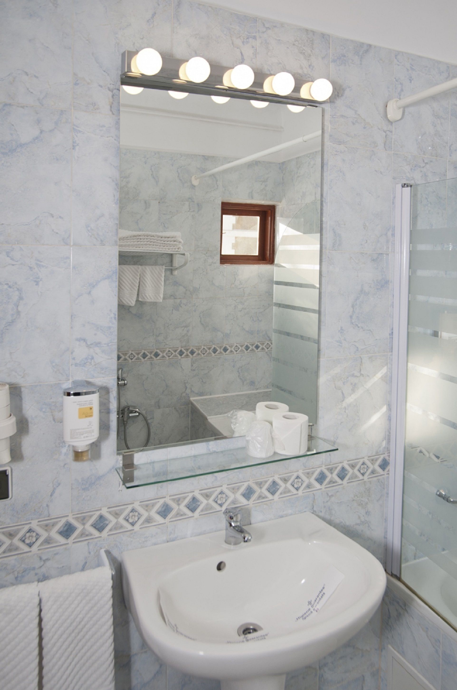Une salle de bain avec un mur recouvert de carreaux de marbre, un lavabo en verre, un miroir avec éclairage intégré, un porte-serviettes et un espace douche.