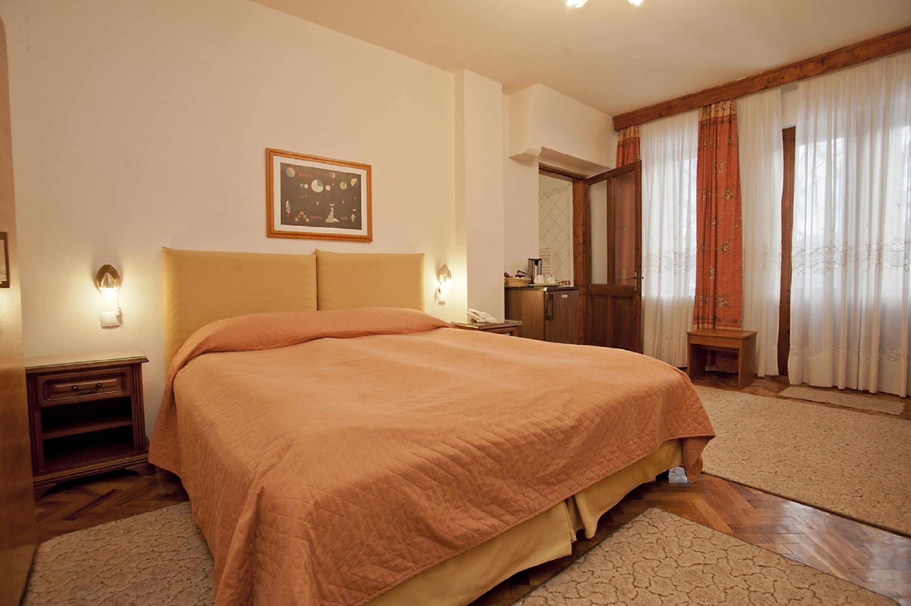 A bedroom with a large bed, wooden headboard, bedside tables, a nightstand, a framed picture on the wall, and a window with curtains.