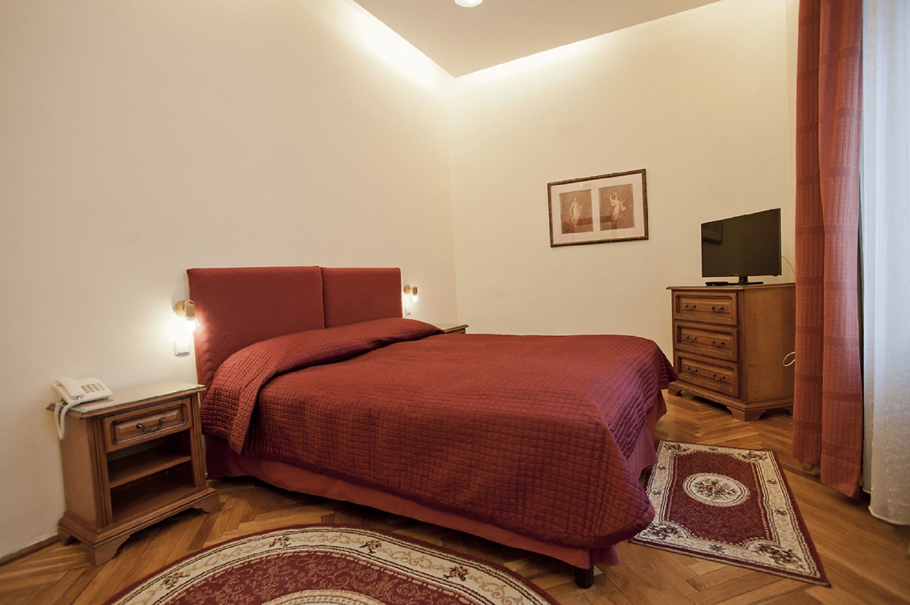 A bedroom with a large bed, a nightstand with a phone, a TV on a wooden cabinet, and a patterned rug.