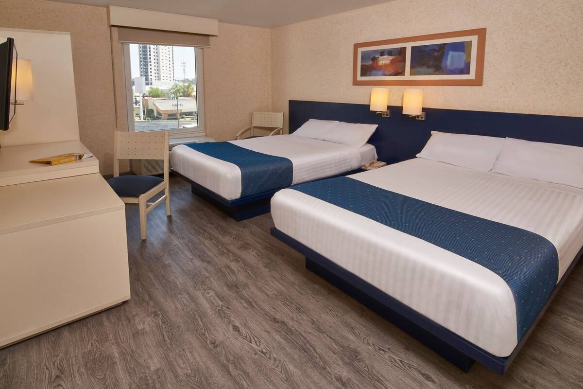 A hotel room with two beds, a desk, a chair, and a window with a view of buildings.