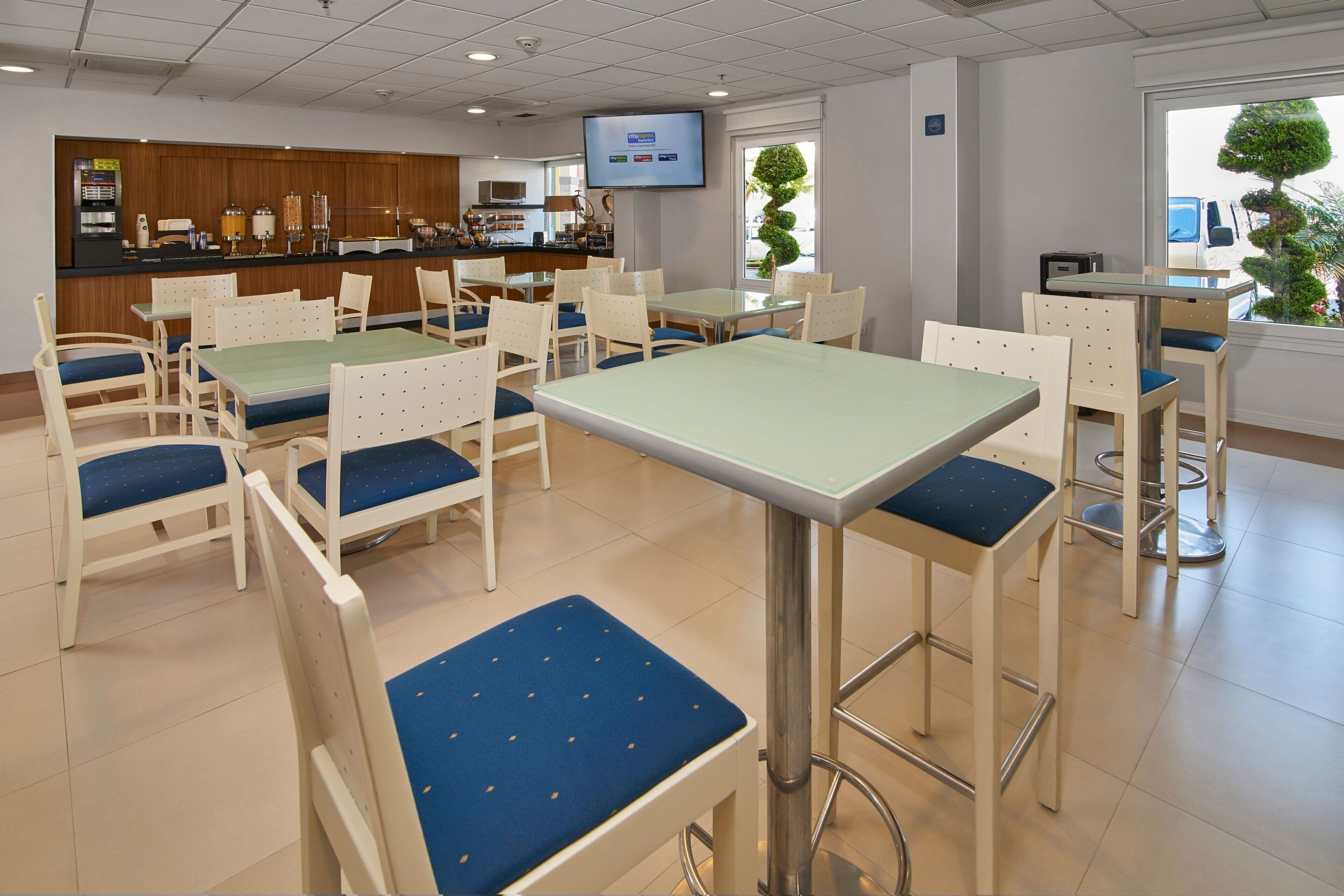 Un comedor de hotel con mesas y sillas, una cocina al fondo y una pantalla de televisión.