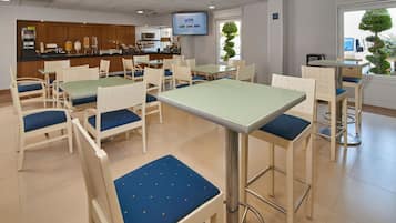 Un comedor de hotel con mesas y sillas, una cocina al fondo y una pantalla de televisión.