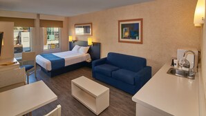 A hotel room with a bed, a blue sofa, a small white table, a kitchenette with a sink, and a window with a view of the outside. - City Express by Marriott Puebla Angelopolis (Puebla)
