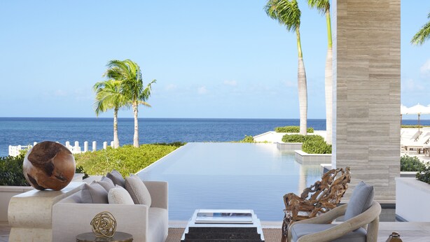 A luxurious outdoor seating area with a view of the ocean, featuring a pool, palm trees, and modern furniture. - Four Seasons Resort and Residences Anguilla (West End Village)