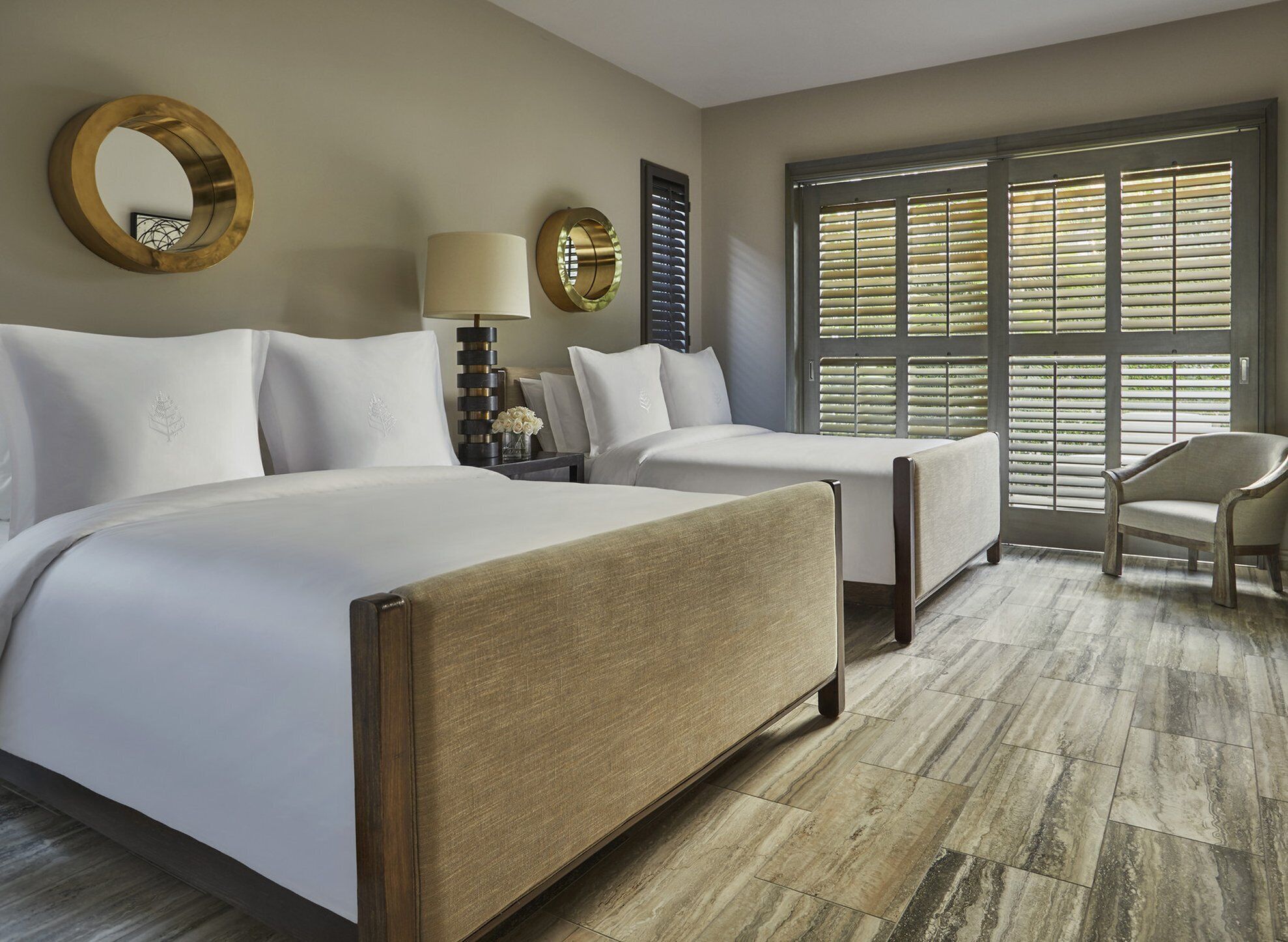 A hotel room with a large bed, two smaller beds, a chair, a mirror, a lamp, and a window with blinds.