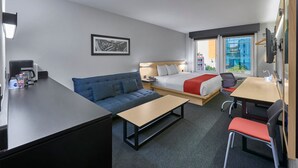 A hotel room with a bed, a sofa, a desk, and a chair. There is a window with a view of buildings outside. - City Express by Marriott Tijuana Río (Tijuana)