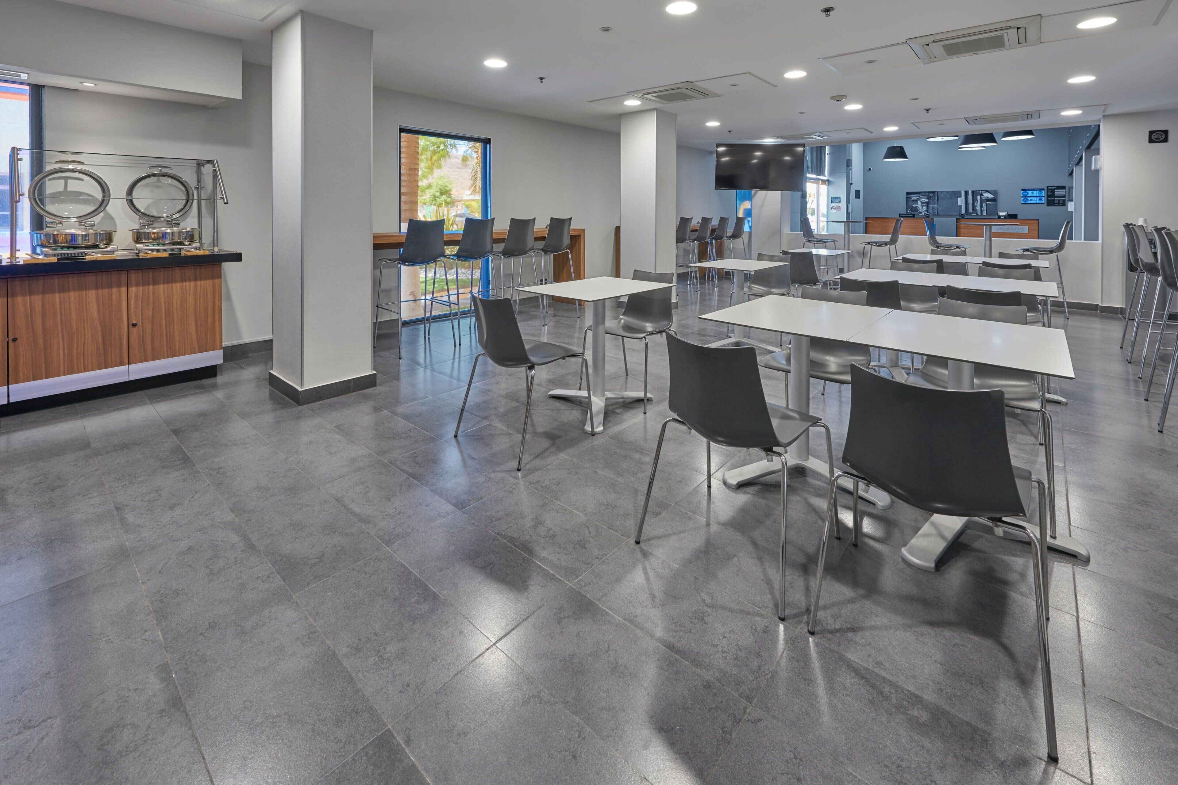 Una cafetería moderna con pisos de baldosas grises, sillas de metal y un mostrador con dispensadores de comida.