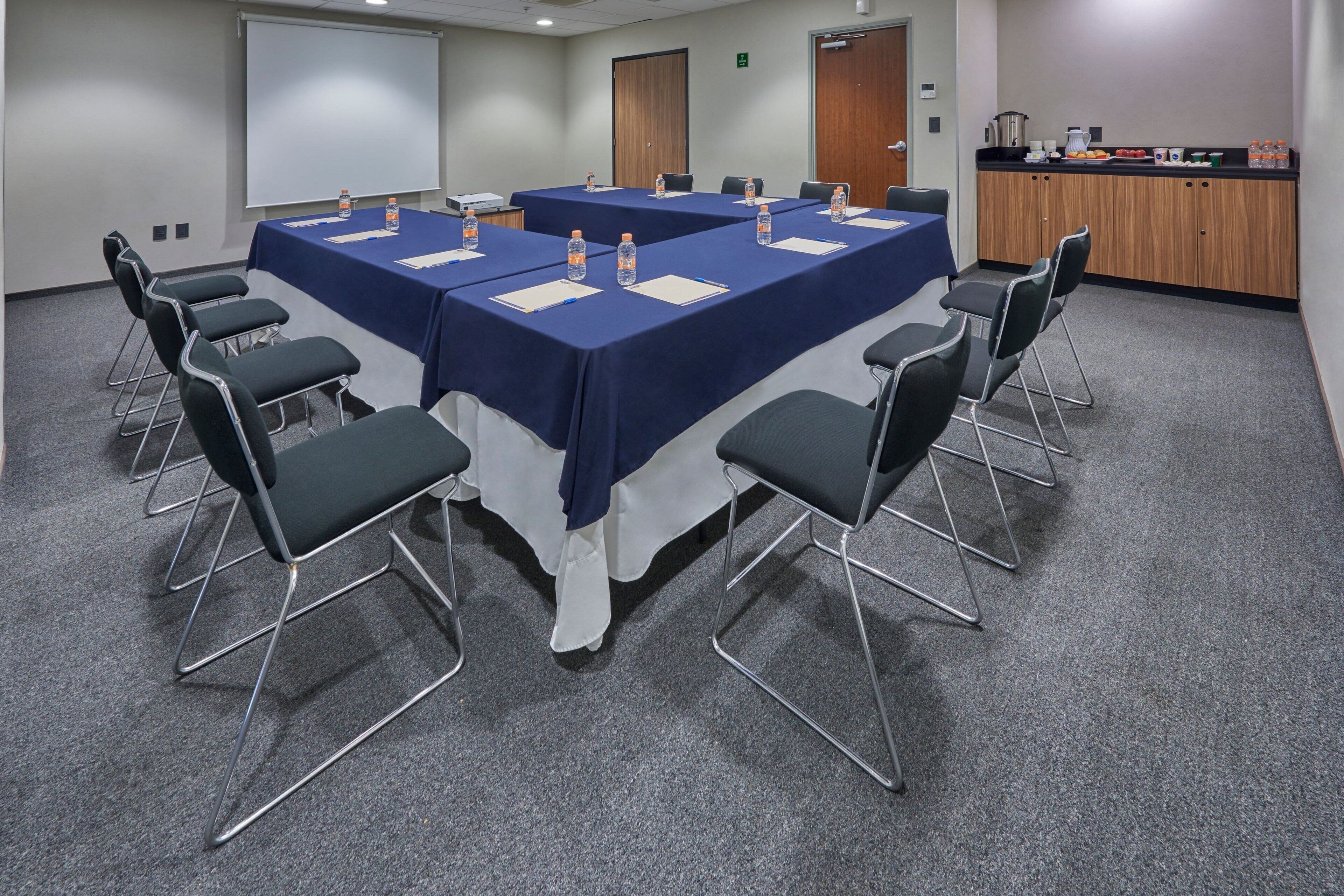 Un salón de conferencias con una larga mesa, sillas, una pantalla de proyector y una zona de kitchenette.