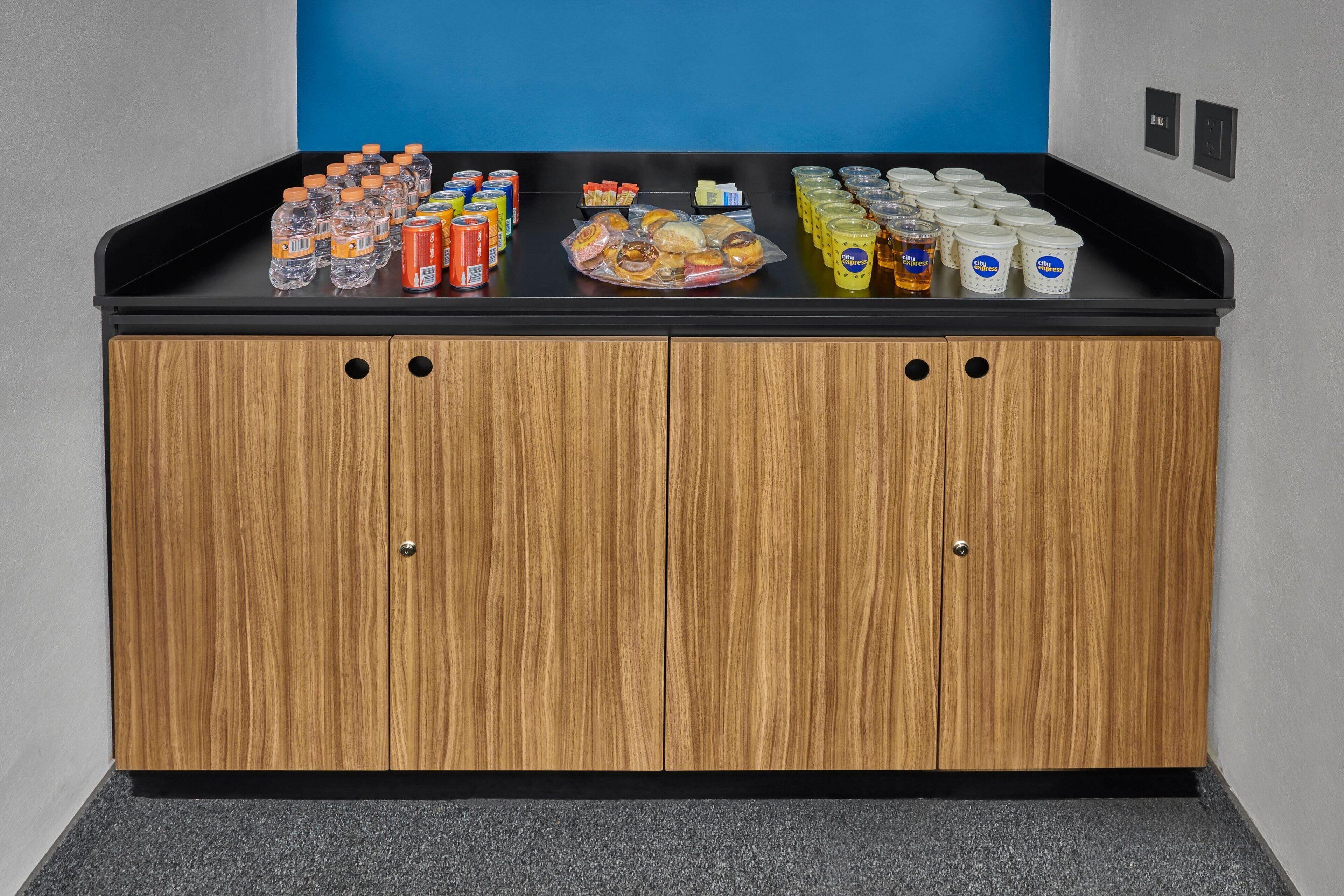 A kitchenette with a black countertop, wooden cabinets, and various food and drink items.