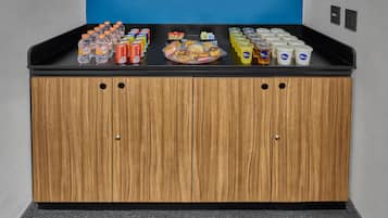 A kitchenette with a black countertop, wooden cabinets, and various food and drink items.