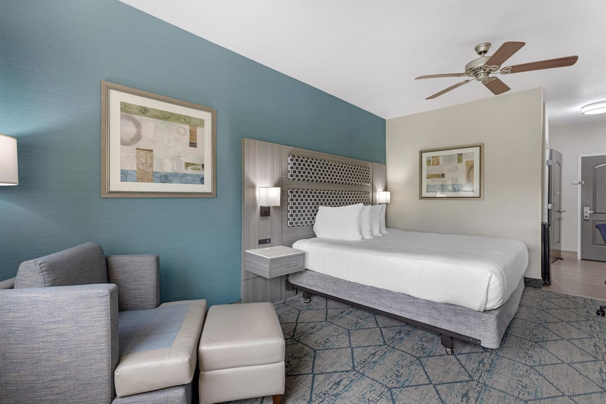 A hotel room with a bed, a seating area with a chair and ottoman, a nightstand, a wall-mounted heater, and a ceiling fan.
