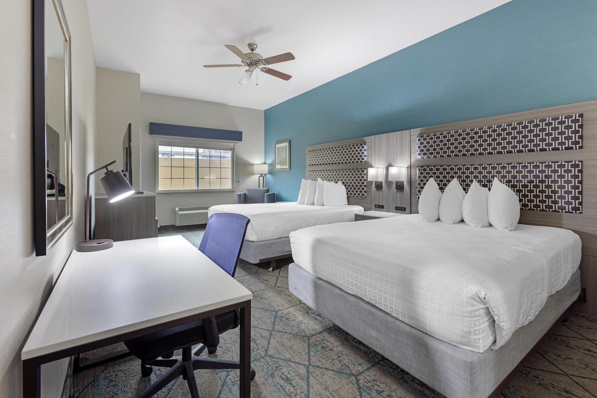 A hotel room with two beds, a desk, a chair, a ceiling fan, and a window.